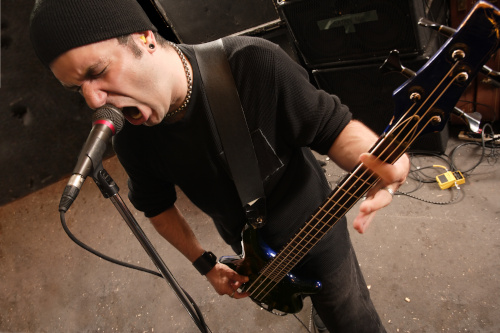 Featured photo for "Heavy Metal Bass Lessons" at Knoxville Bass Lessons depicting man in black clothing and black skull cap with extremely low-slung bass guitar screaming into a microphone.
